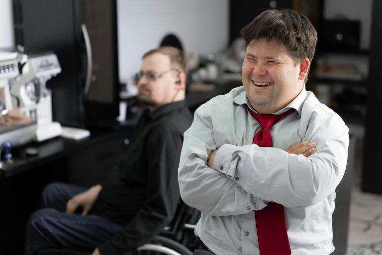 Un homme en situation de handicap sourit. Il est sur son lieu de travail, un ESAT. On voit un homme en fauteuil roulant en arrière-plan.