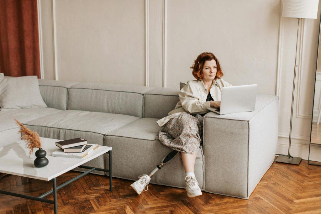 une femme avec une prothèse à la jambe gauche est assise sur un canapé et travaille sur son ordinateur portable, posé sur l'accoudoir du canapé.